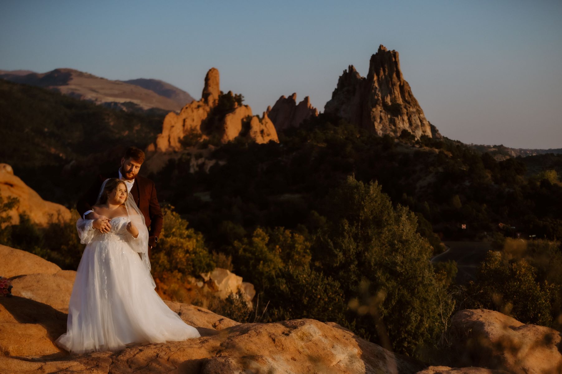 A couple standing on top of rocks and behind them are larger rock formations they are standing together and one is wearing a white wedding dress and white veil and looking off to the side the other is wearing a suit and looking down at them