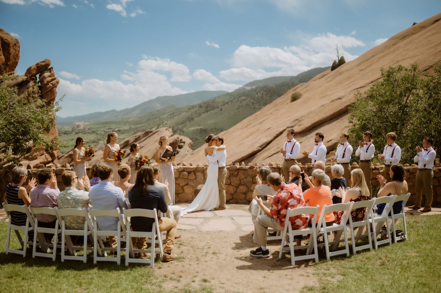 A wedding ceremony with guests and a wedding party. The guests and wedding party and clapping for the couple and the wedding ceremony takes place outdoors. The couple is embracing. 