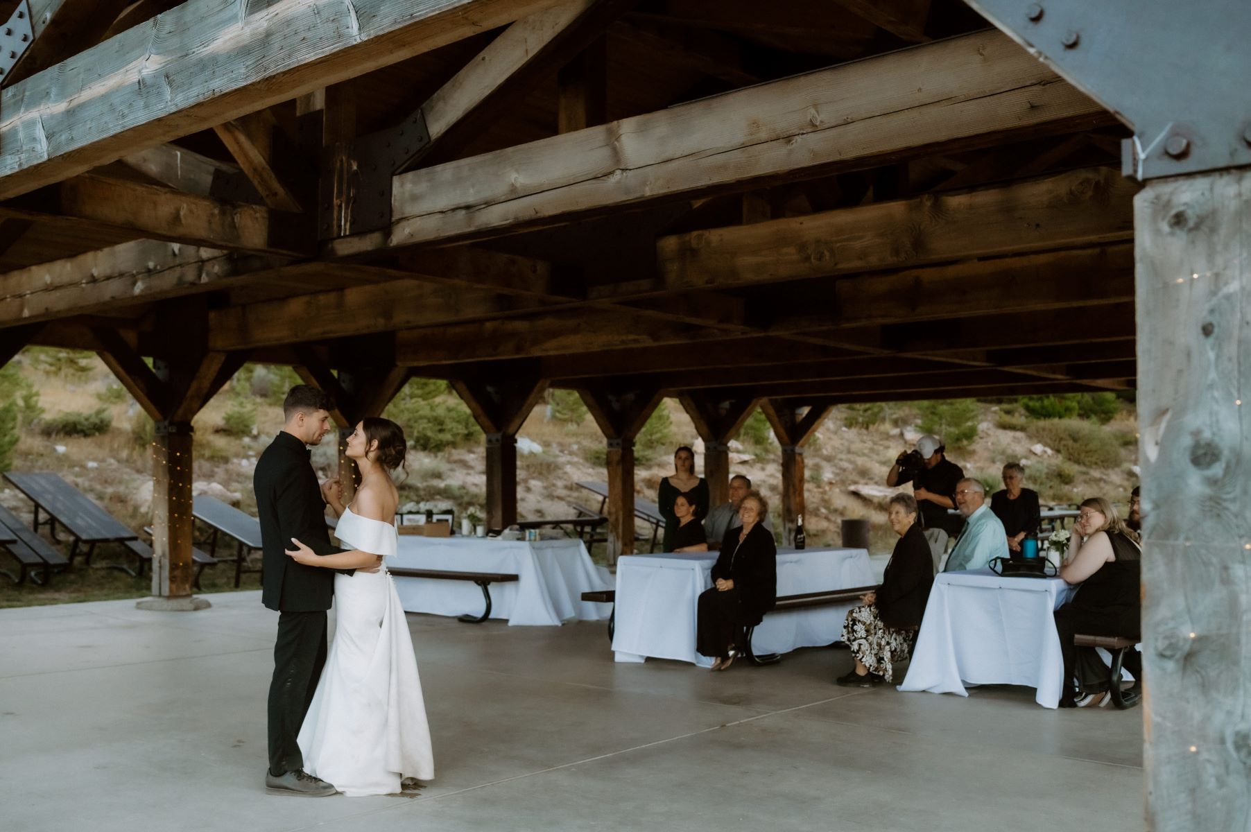 A couple dancing at their wedding reception as their guests sit at picnic tables covered in white cloths and watch them 