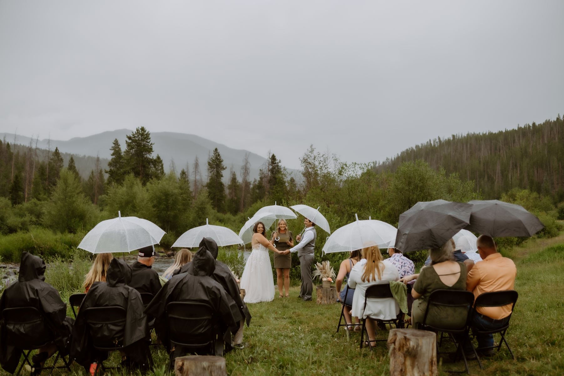 A couple at their wedding ceremony holding hands with their officiant behind them are mountains and evergreen tress and their guests are seated in chairs in front of them also everyone is using white and black umbrellas 