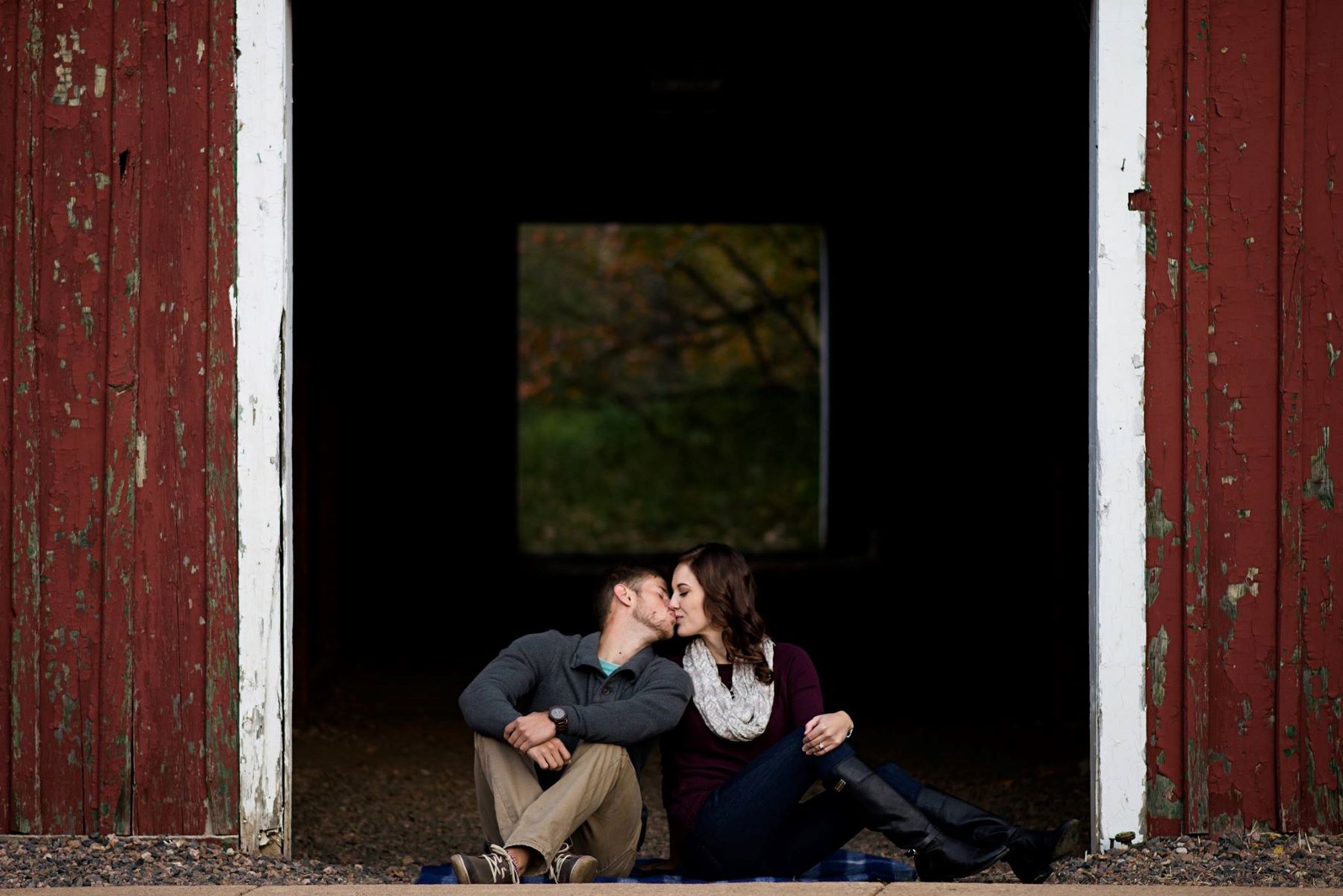 A couple kissing each other and sitting down leaning towards each other in front of a large red barn