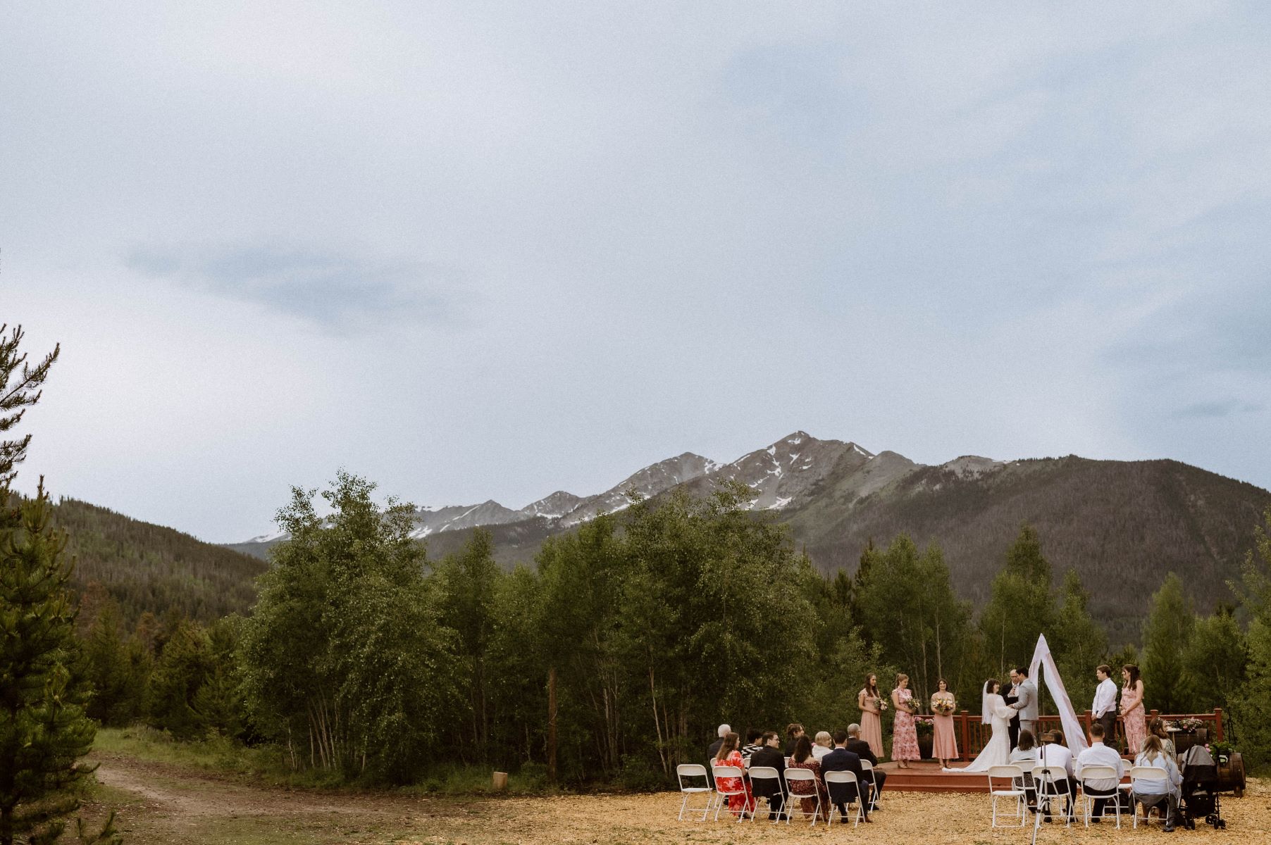 A couple at their wedding ceremony with their wedding officiant and the mountains as their backdrop their wedding party is next to them and their weddding guests are sitting in front of them 