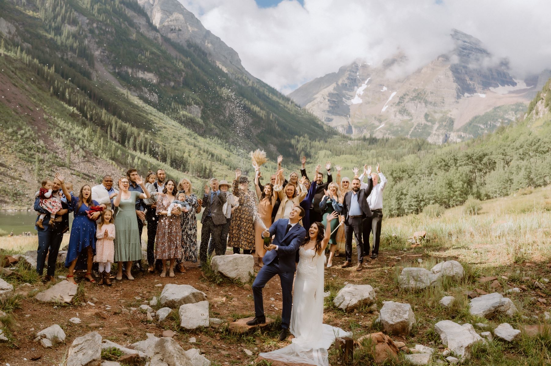 A couple celebrating their marriage the groom is popping a bottle of champagne and behind them is their wedding party celebrating and the mountains