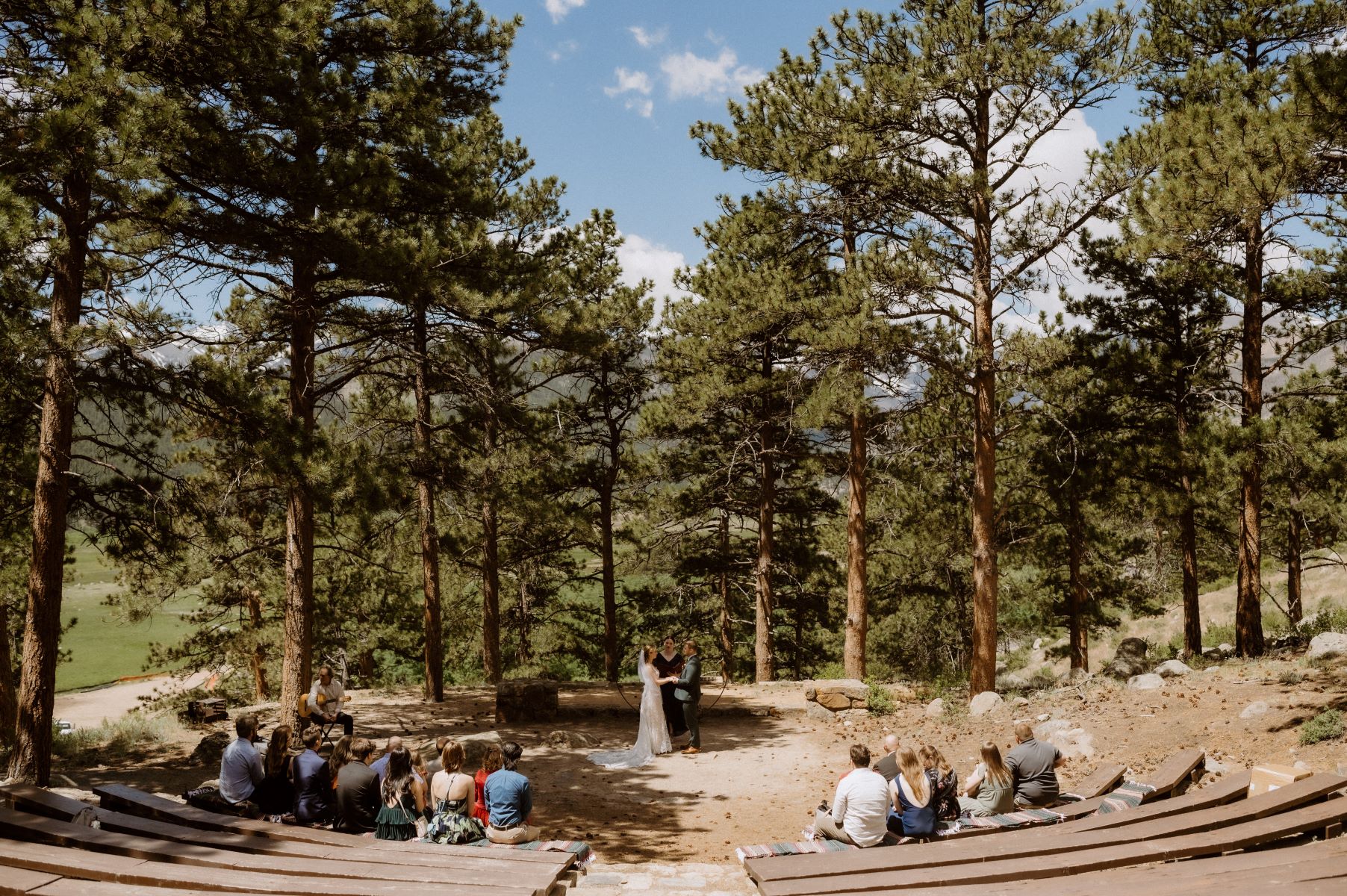 A couple at their wedding ceremony with their officiant surrounded by trees and their guests are sitting in front of them