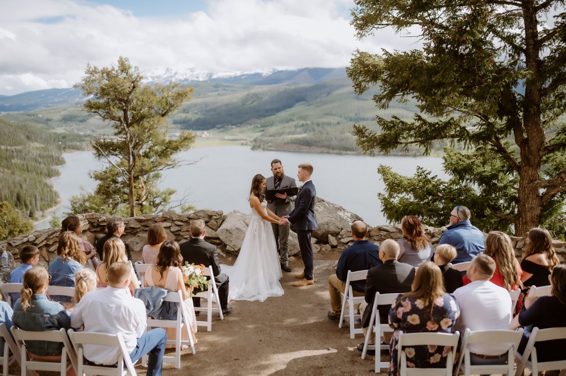 A couple at their wedding ceremony with their officiant behind them is a lake and mountains the couple is holding hands and the groom is looking at his partner the wedding guests are sitting in chairs watching them 