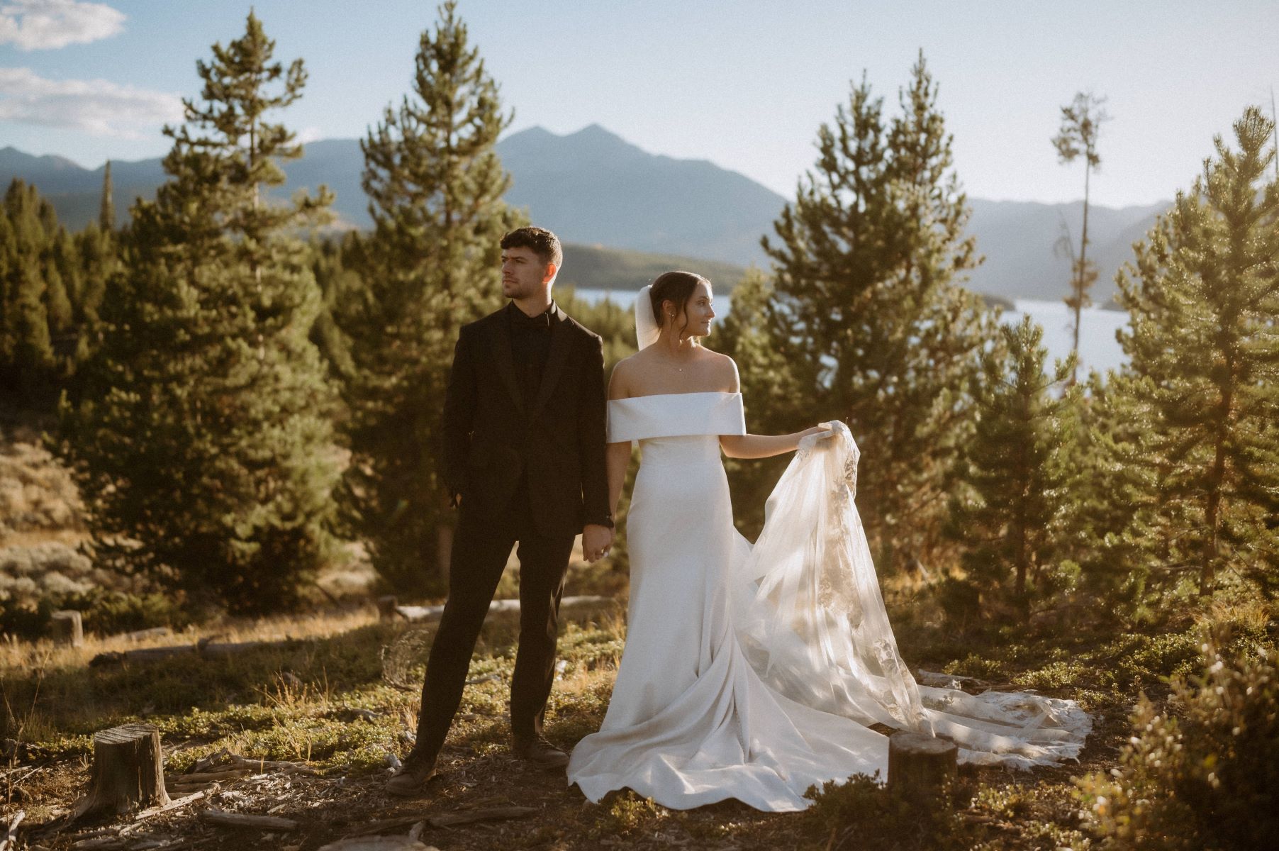 A couple standing together and holding hands behind thema re mountains and a lake and evergreen treess the woman is wearing a wedding dress the man is wearing a black suit 