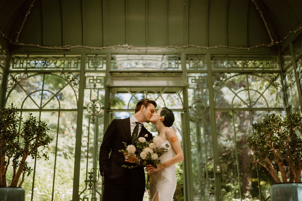 A newlywed couple kissing each other as one holds a bouquet of flowers 