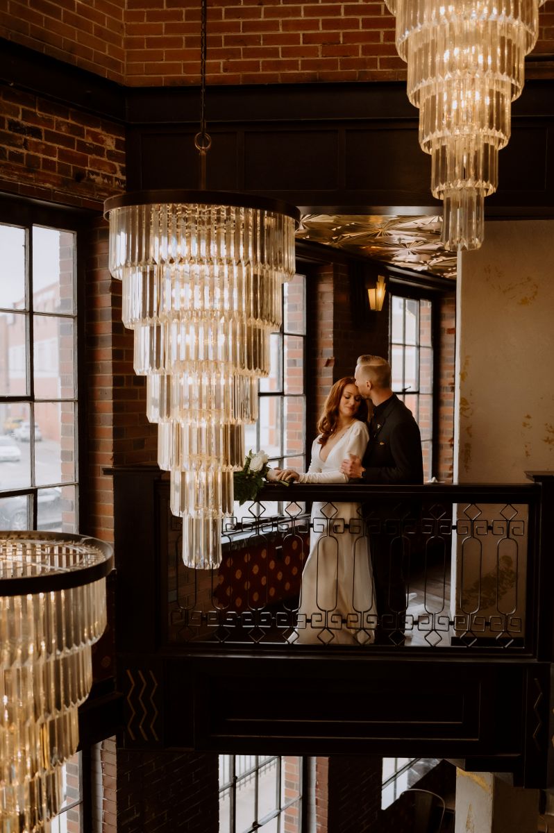 a couple together next to a blackrailing in front of them are large chandeliers the man is kissing his partner on the head and holding her arm with one hand the woman is wearing a white wedding dress and holding a bouquet of flowers 