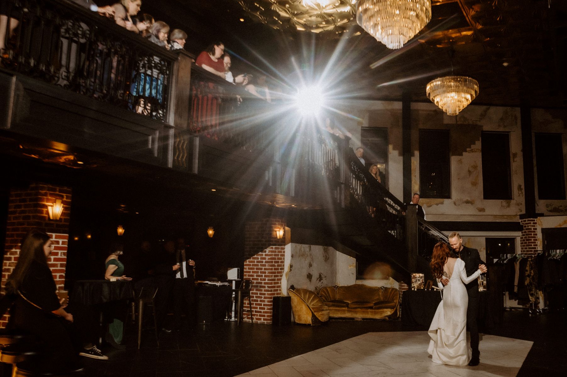 a couple at their wedding dancing together ont he dance floor as their guests watch the woman is wearing a white wedding dress the man is wearing a black suit some guests are standing on the staircase and watching the couple dance above everyone's heads are large chandeliers 