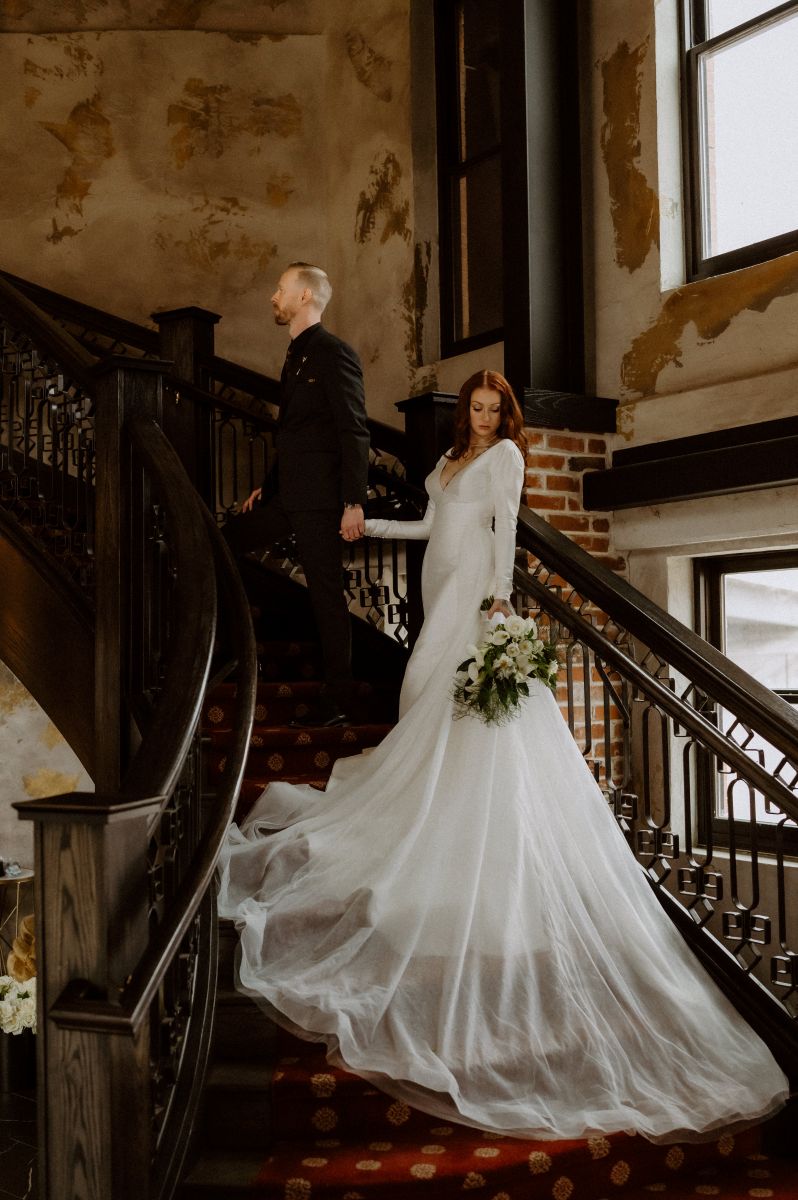 a couple on a grand staircase the man is wearing a black suit the woman is wearing a white wedding dress and looking down towards the bottom of the stairs she is also holding a bouquet of flowers 