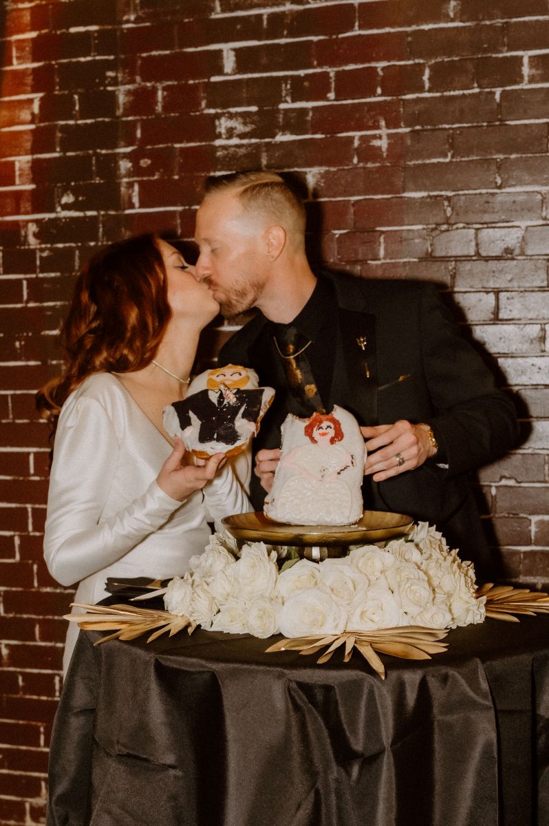 a couple at their weddigng reception next to their wedding cookies they are kissing one another 