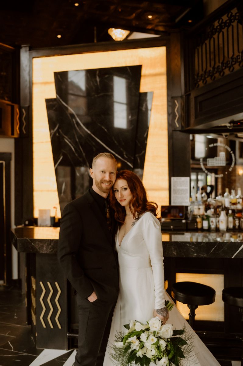 a couple standing together in front of a bar the woman is wearing a white wedding dress and holding a bouquet of flowers the man is wearing a black suit 