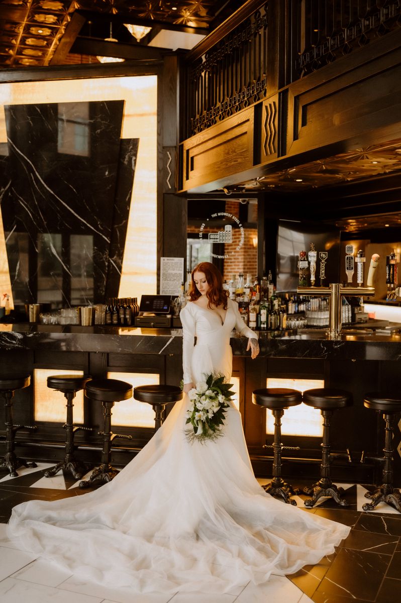 a woman in a white wedding dress holding a bouquet of flowers and leaning against a bar