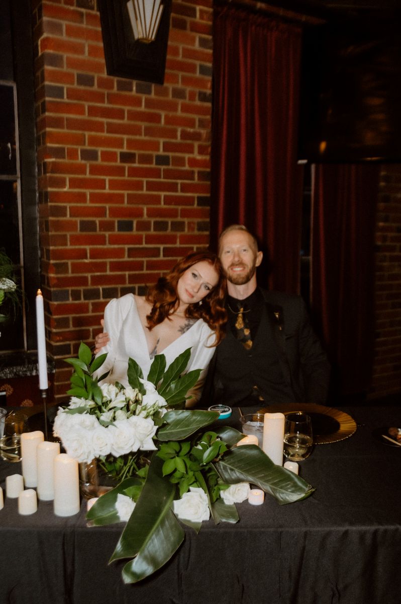 a couple sitting at a table together the man is putting his arm around his partner woh is wearing a white wedding dress in front of them are candles and plants and flowers 