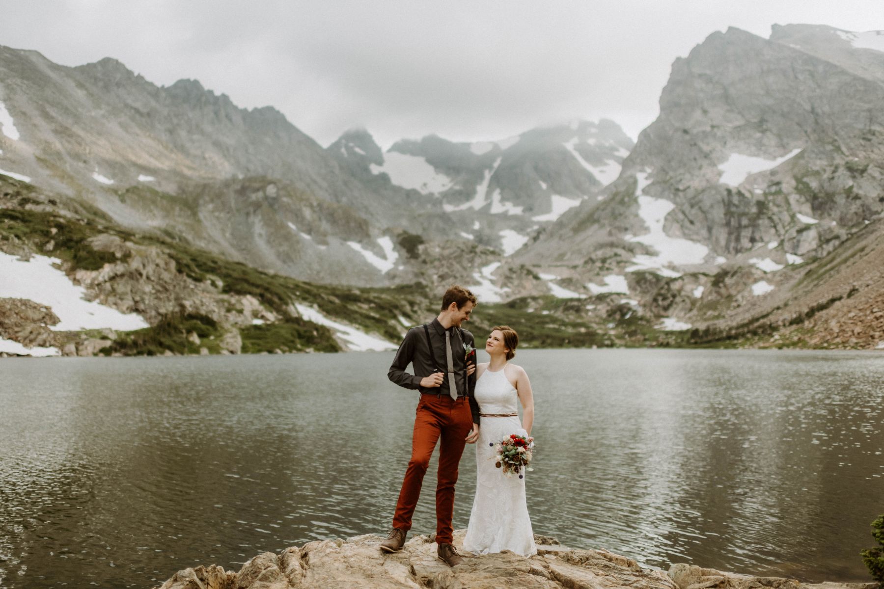 a copule in front of a lake standing next to each other and looking at one another behind the lake are mountains covered in snow the woman is wearing a white dress and holding flowers 