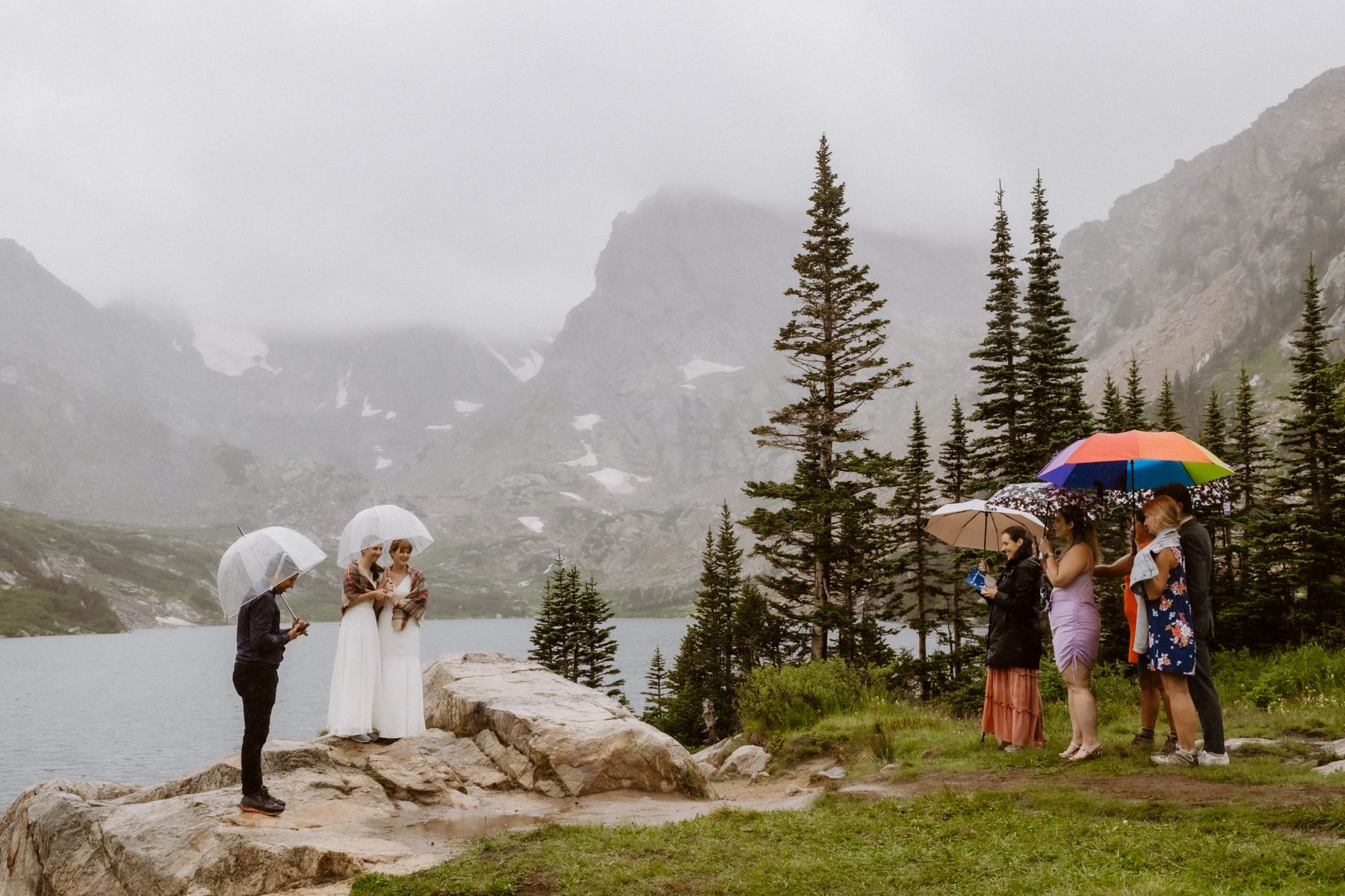 two women having their wedding ceremony at lake isabelle in colorado it is raining so they both have umbrellas and so do their guests 