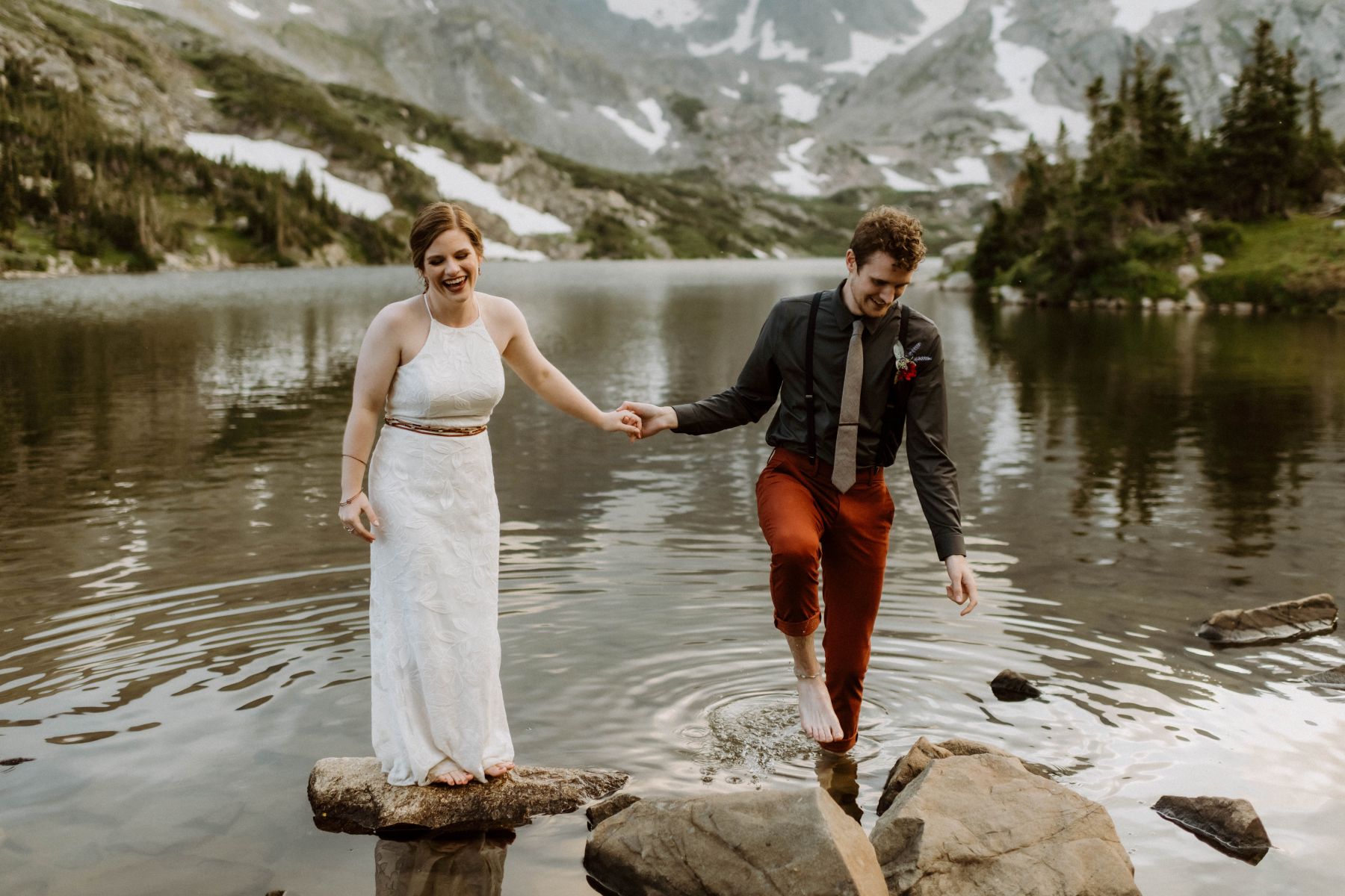 a couple together at a lake the woman is standing on top of a rock and laughing the man is walking through the water and holding her hand 