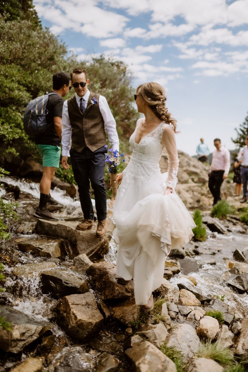 a woman wearing a white wedding dress and sunglasses is holding a bouquet of flowers and looking back to her partner who is wearing a white button up , pants, and a dress vest 