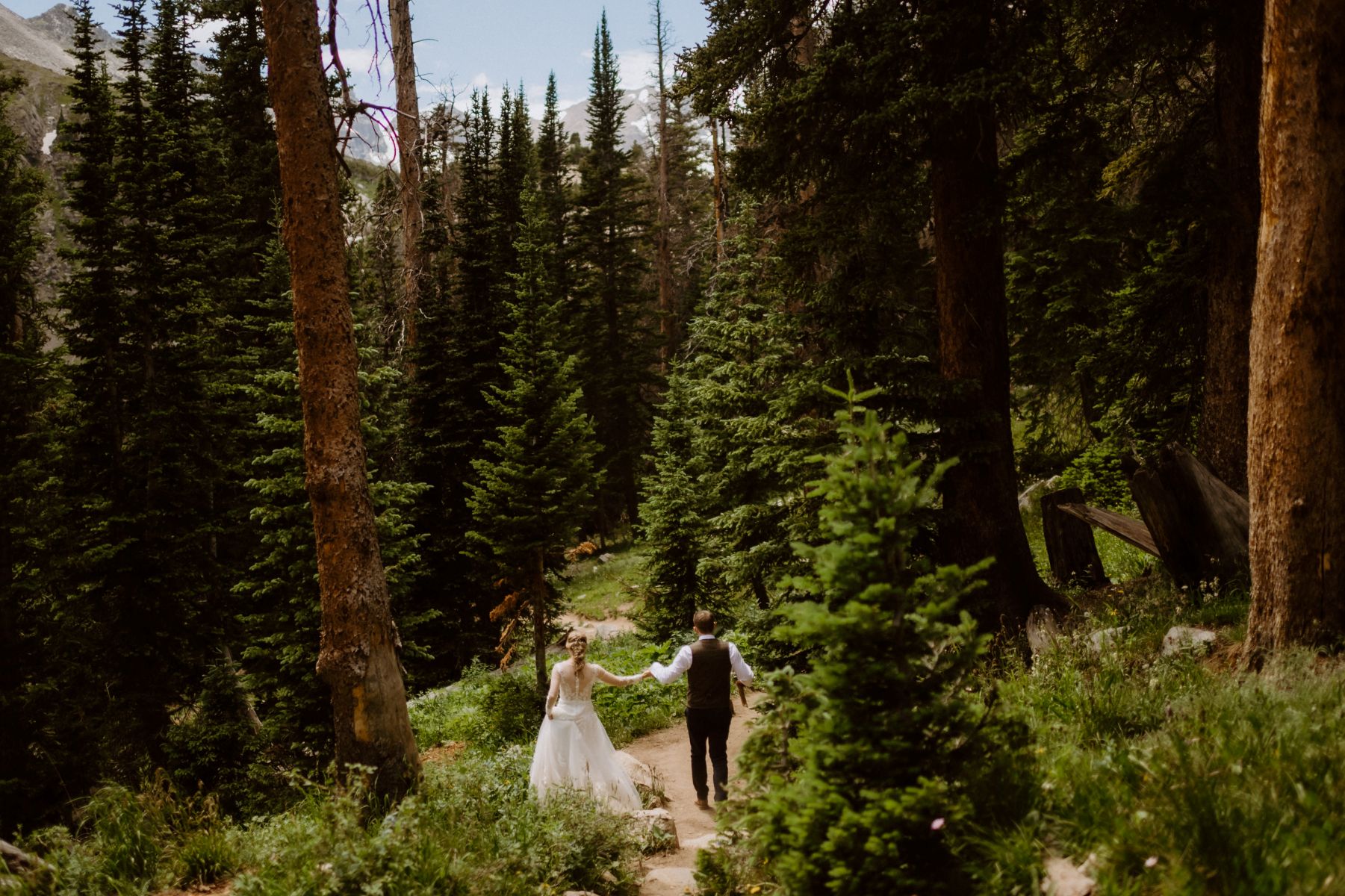 a couple walking hand ni hand through a forest filled with pines the woman is wearing a white wedding dress the man is wearing a white button up , pants, and a dress vest 