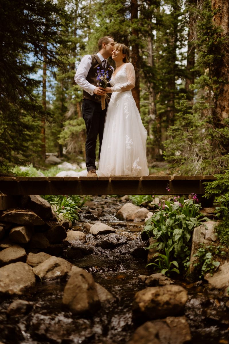 a couple standing on a wooden plank in the forest the man is kissing the woman's cheek the woman is wearing a white wedding dress and smiling and she is holding a bouquet of flowers 