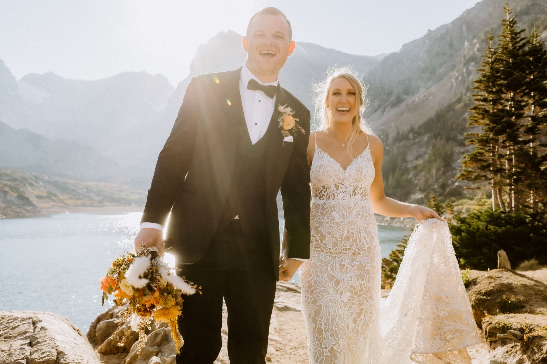 a couple on top of rocks next to a lake the woman is wearing a white wedding dress the man is wearing a black suit and holding flowers both are smiling 
