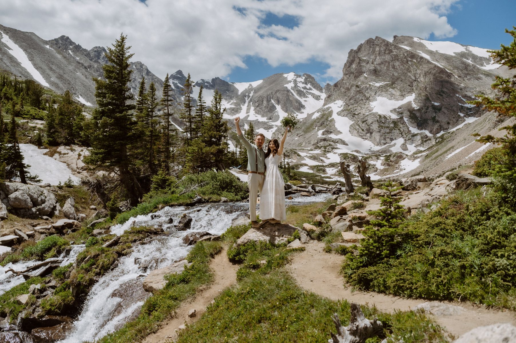a couple next to a waterfall holding their arms up in the air behind them are pines and mountains 