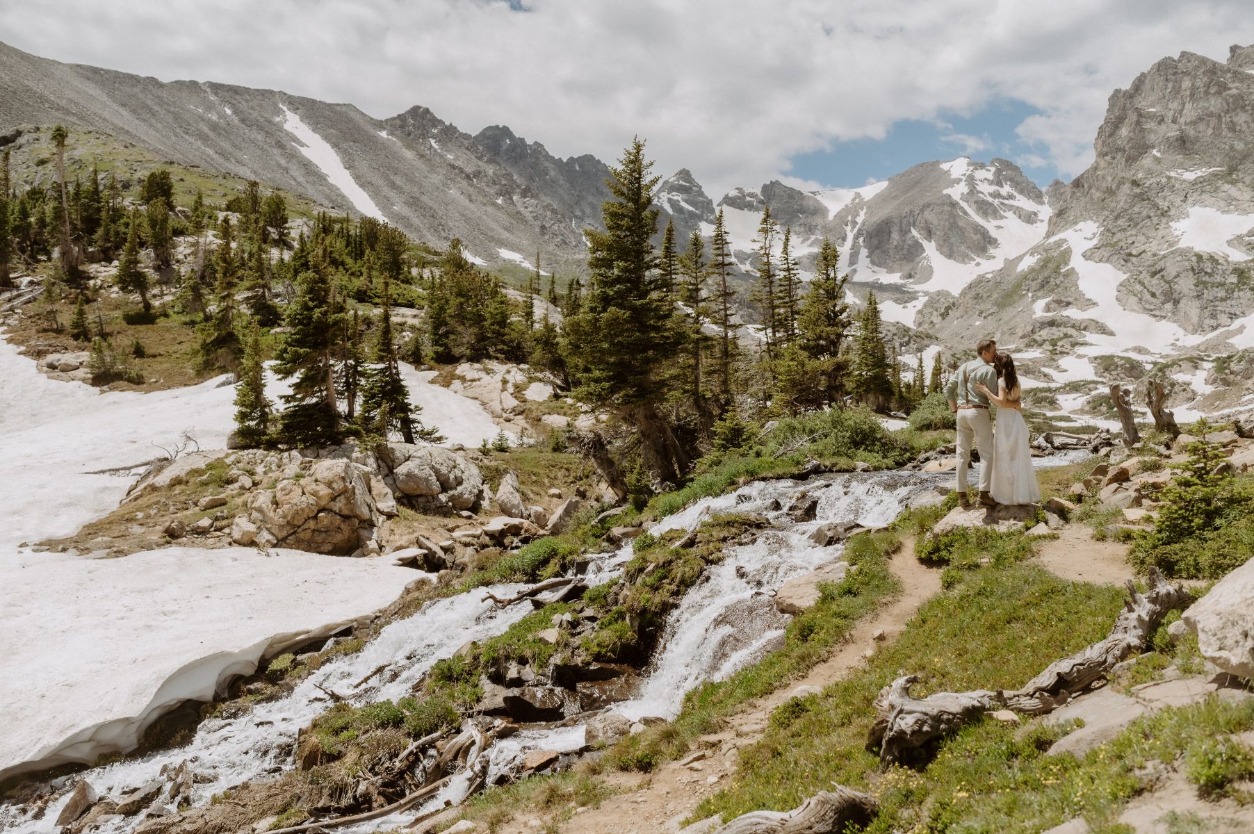 a couple standing next to a waterfall and looking at the mountains behind them that are covered in snow and there are pines too 