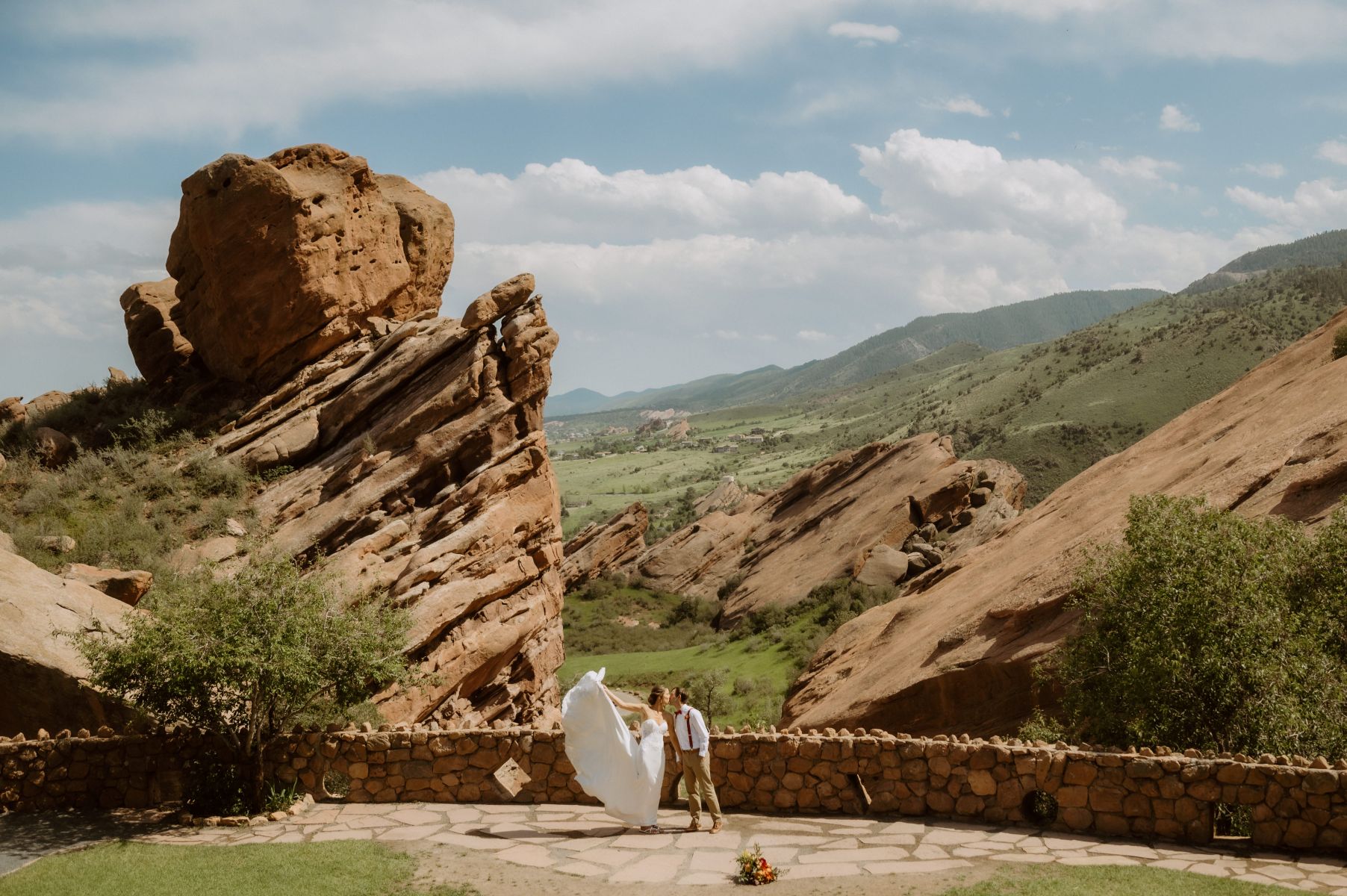 a couple kissing each other at red rocks trading post the bride is lifting her dress up behind her and behind them are views of red rocks formations and a valley and mountains 