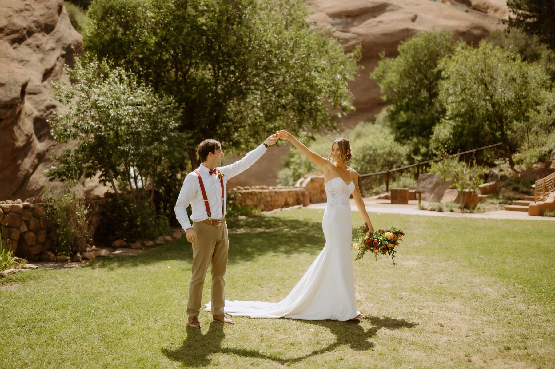 a couple on a patch of grass holding their hands up in the air and looking at each other and smiling the woman is wearing a white wedding dress and holding a bouquet of flwoers the man is wearing a white button up and tan pants