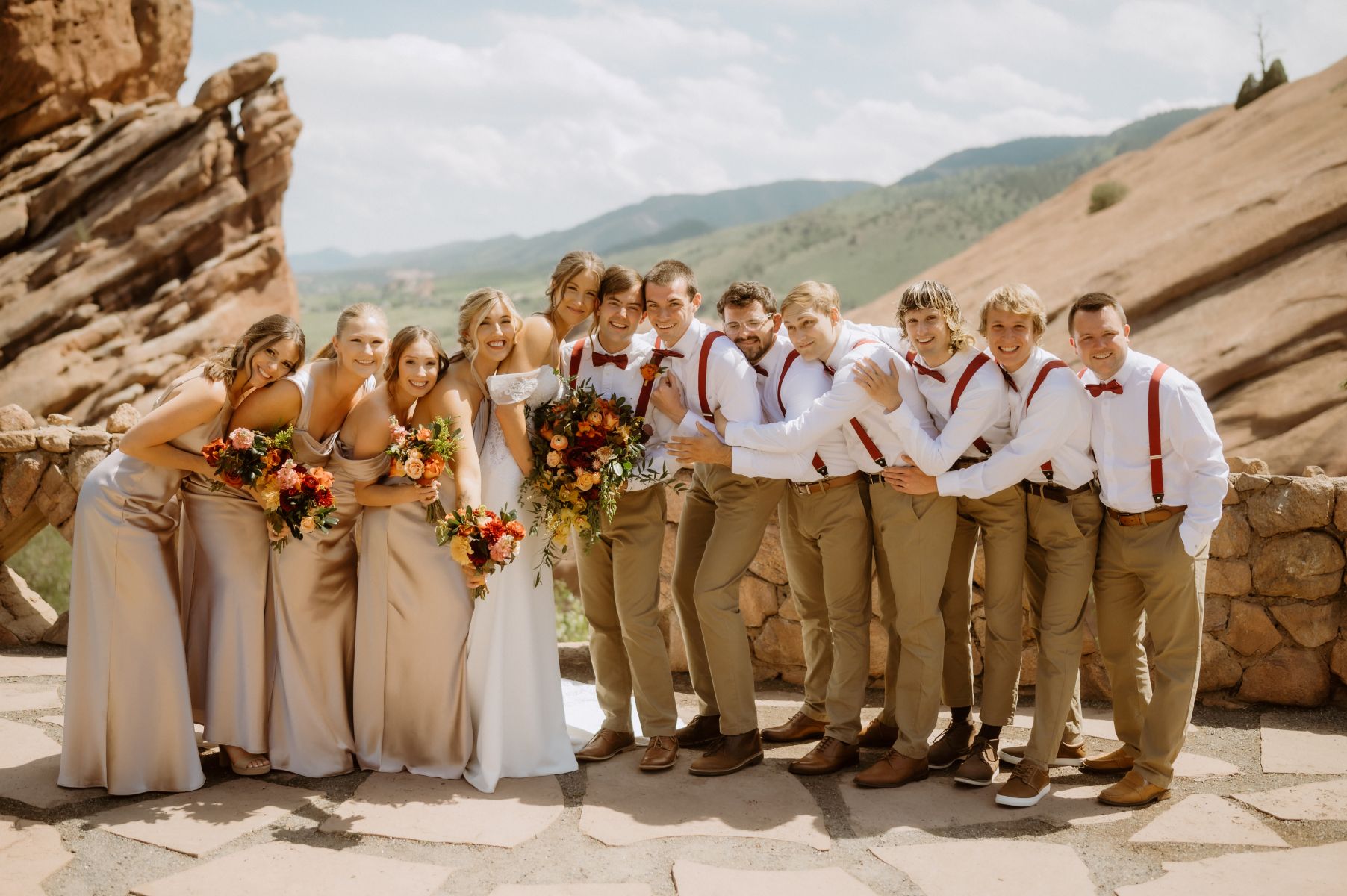 a couple with their wedding party they are all smiling and leaning into each other the men are wearing white button ups and tan pants with red suspender straps the women are wearing tan dresses and the bride is wearing a white wedding dress and holding a bouquet of flowers 
