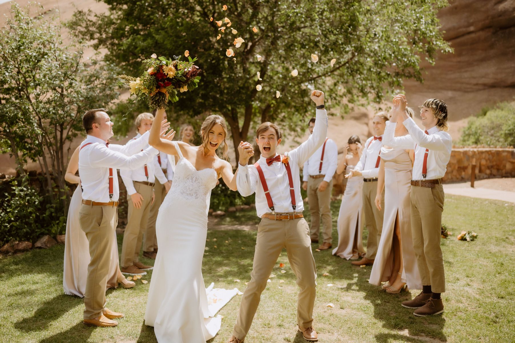a couple cheering and holding their hands in the air and smiling behind them their wedding party is throwing wedding petals in the air and cheering for them 