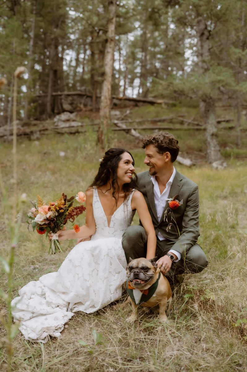a couple in the woods kneeling down and smiling at each other they are both holding onto the dog they are both smiling and laughing with each other the woman is wearing a white wedding dress the man is wearing a green suit 
