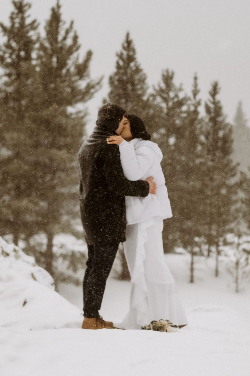 a couple kissing in the snow surrounded by pines tress the woman is wearing a white wedding dress and jacket the man is wearing a brown jacket and earmuffs 