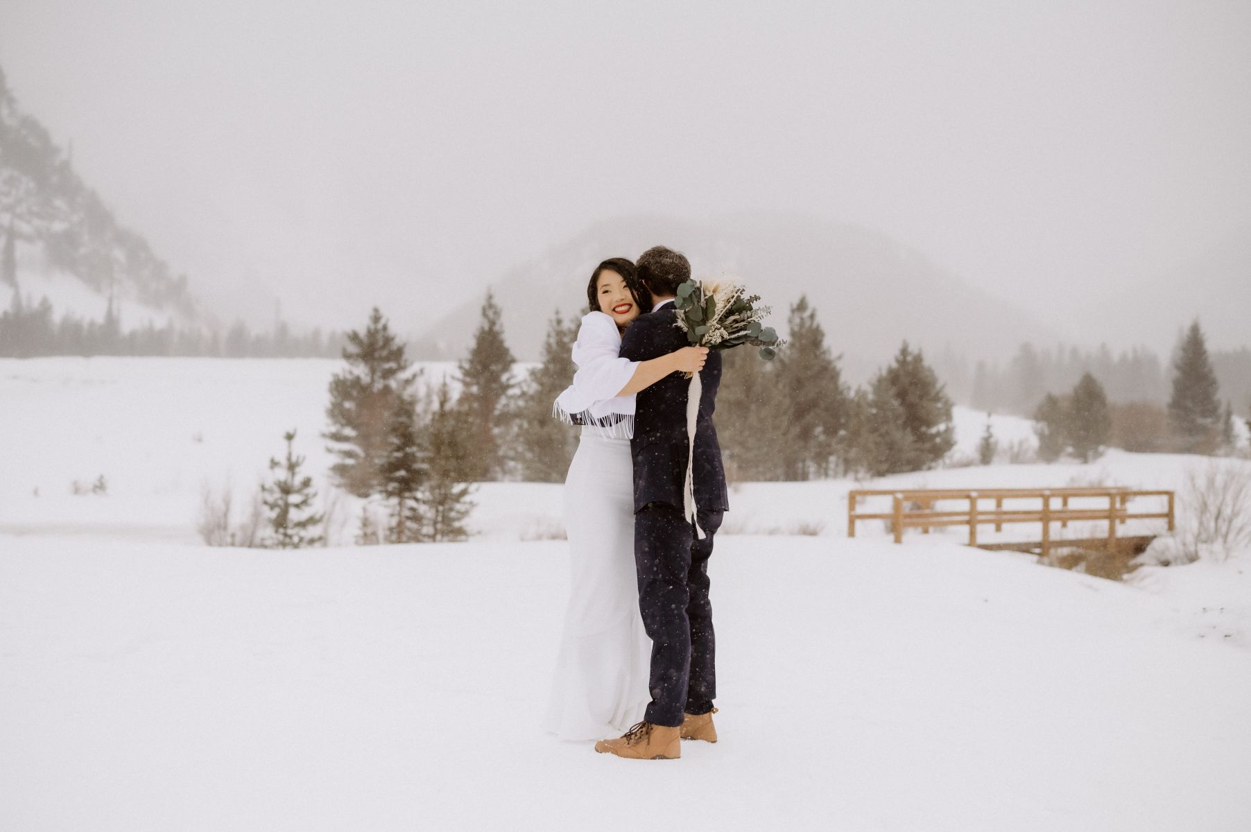 a couple hugging in the snow surrounded by mountains and pines trees 