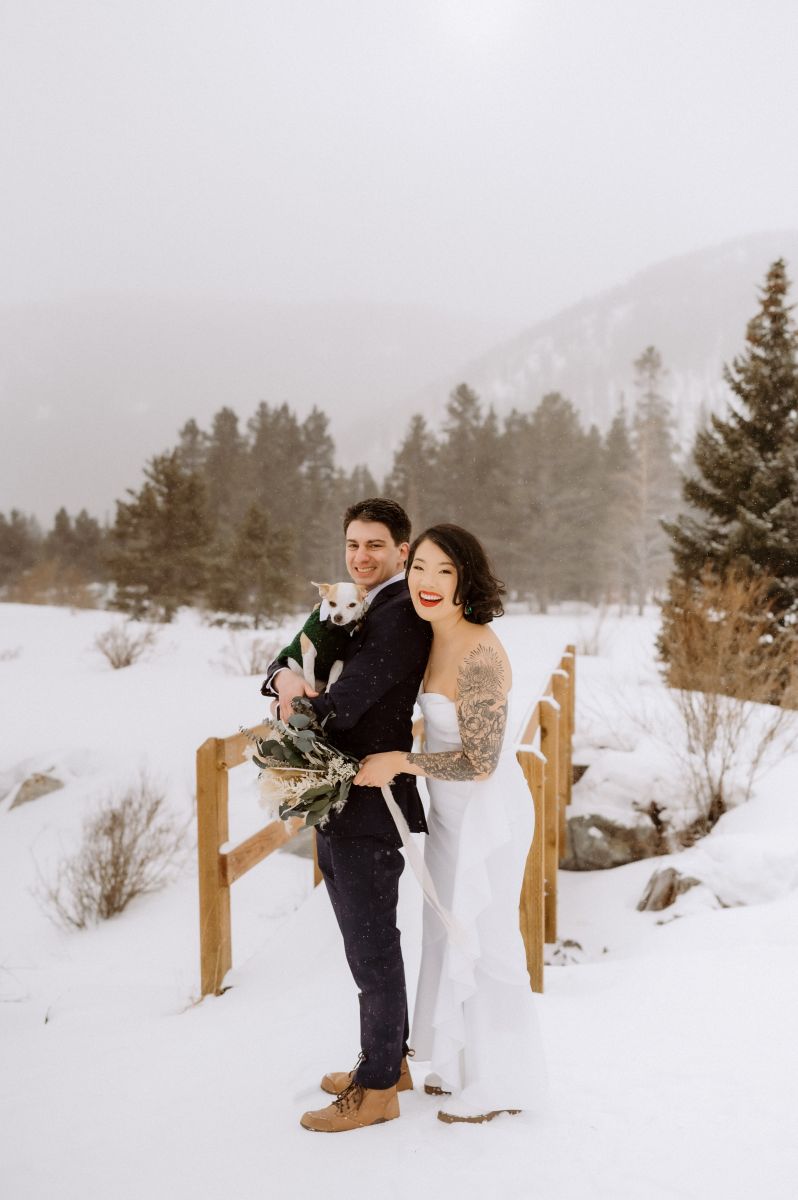 a couple satnding in the snow with their dog the woman is wearing a white wedding dress and holding a bouquet of flowers the man is wearing a black suit and holding a dog behind them are mountains and a snow covered bridge 