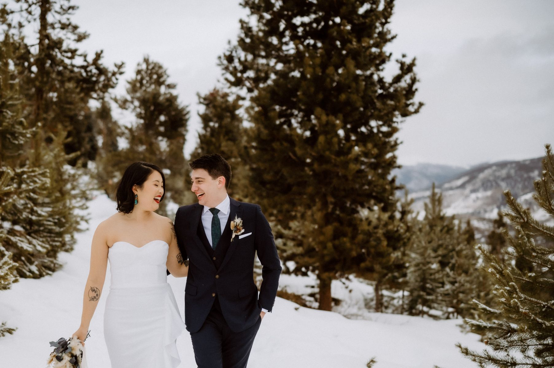 a couple laughing together and embracing as they walk through the snow while surrounded by pine trees the woman is wearing a white wedding dress and holding flowers the man is wearing a black suit 