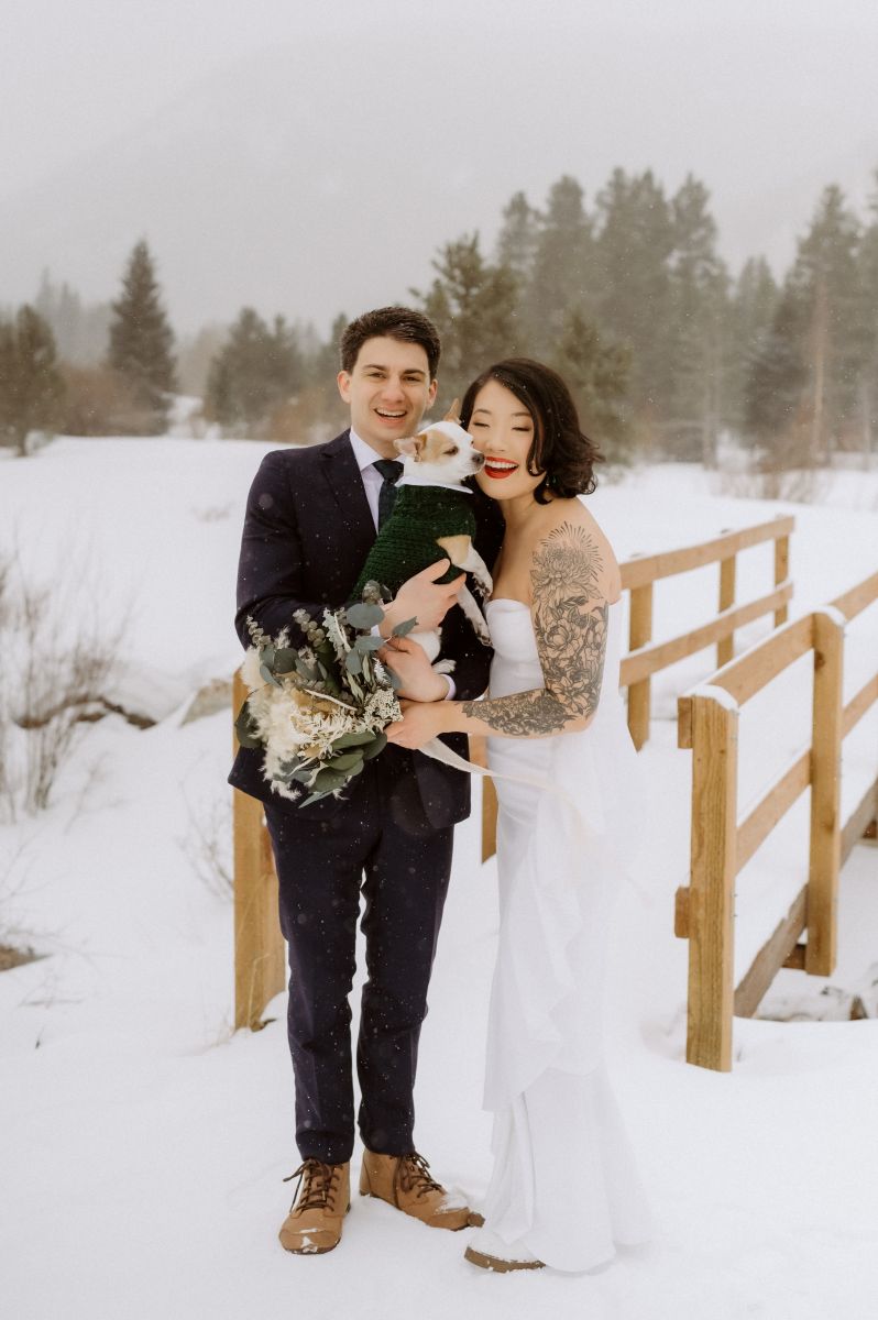 a copule smiling and holding their dog in front of a snow covered bridge in Officer's Gulch in Colorado the woman is wearing a white wedding dress and holding a bouquet of flowers the man is wearing a tuxedo and holding their dog 