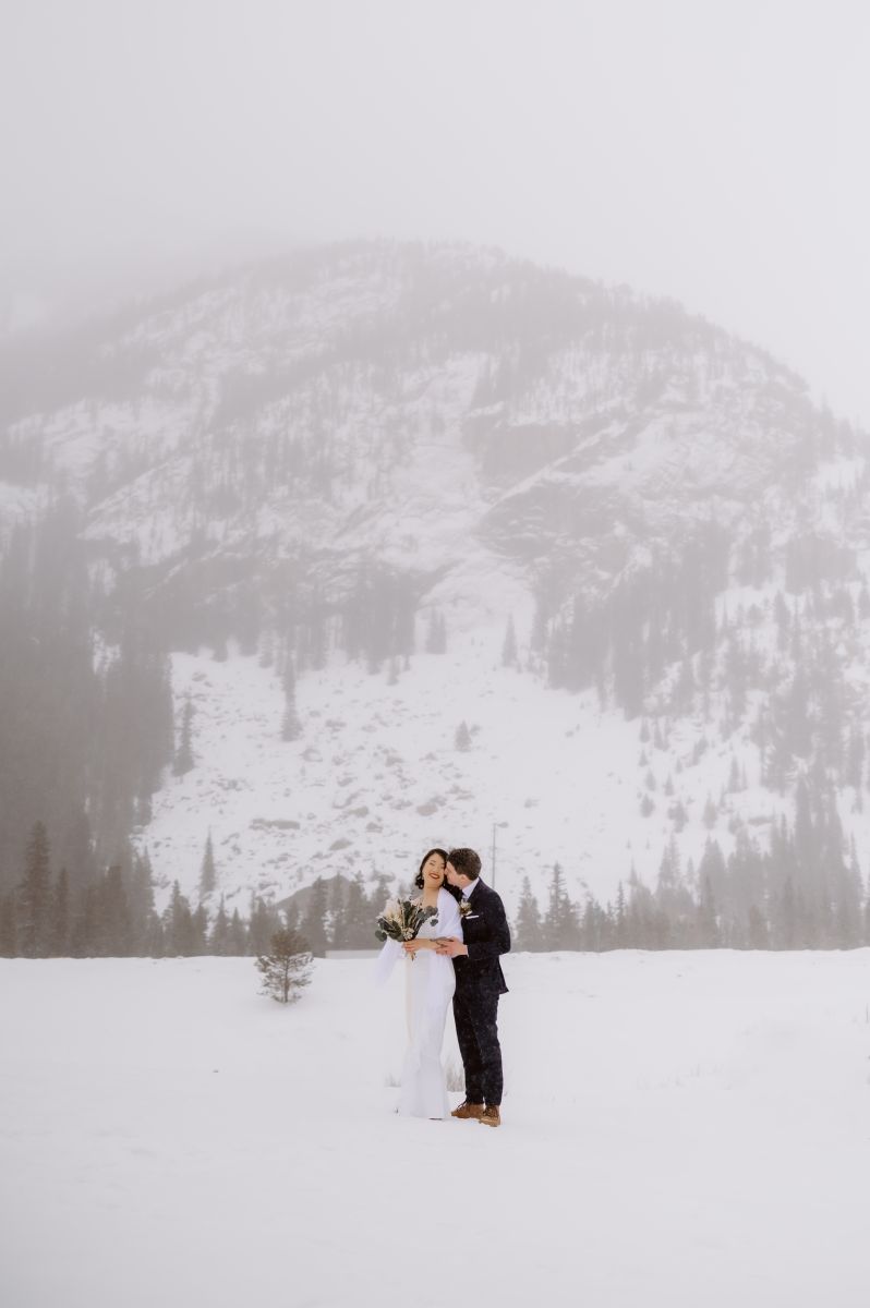 a man kissing his partner's cheek in the snow in front of a mountain the woman is wearing a white wedding dress and blanket the man is wearing a black suit 