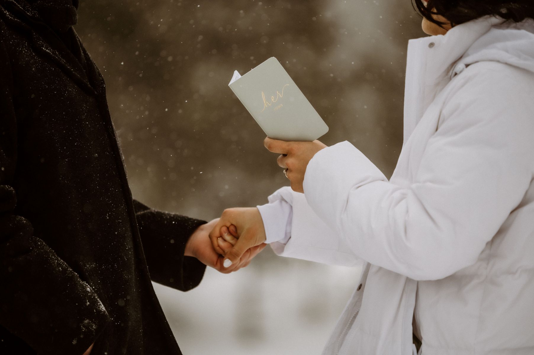 a couple sharing their vows at their winter elopement it is snowing and the woman is wearing a white jacket and holding a vow book and holding her partner's hand 