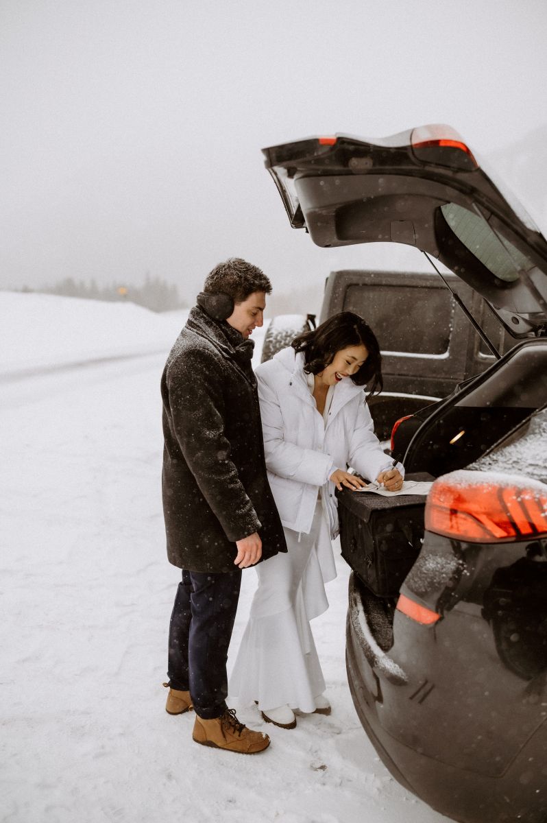 a couple signing their marriage certificate in the trunk of a car laughing together as it snows around them the woman is wearing a white jacket and wedding dress the man is wearing a brown jacket and earmuffs 