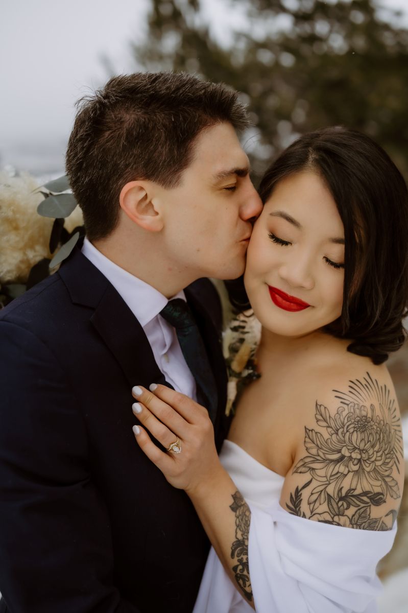 a man kissing the side of his partner's cheek the woman is wearing a white wedding dress and is smiling and her eyes are closed 