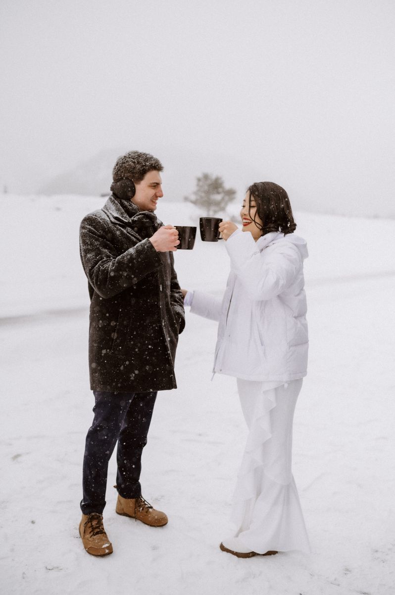 a couple surrounded by the snow and mountains drinking coffee from coffee mugs the woman is wearing a white jacket the man is wearing a brown jacket and ear muffs 