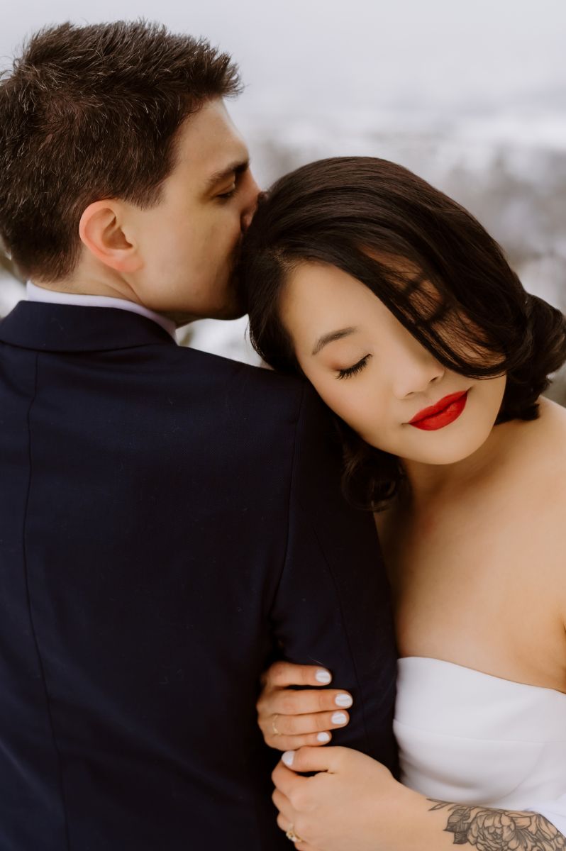 a man kissing the side of his partner's head the woman is smiling and has her eyes closed she is wearing a white wedding dress