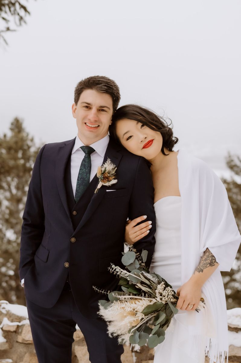 a woman laying her head against the shoulder of her partner and holding his arm at Sapphire Point in Colorado in the winter the man is wearing a black suit and the woman is wearing a white wedding dress and white blanket and holding a bouquet of flowers 