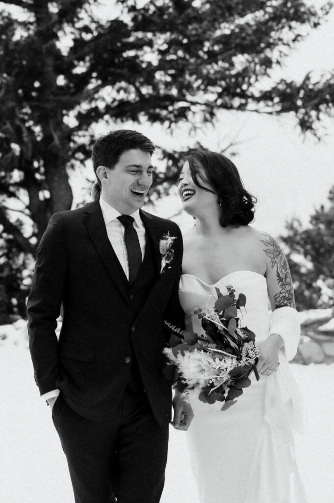 a couple laughing together and walking thorugh the snow the woman is wearing a white wedding dress and white blanket and holding a bouquet of flowers the man is wearing a black suit 