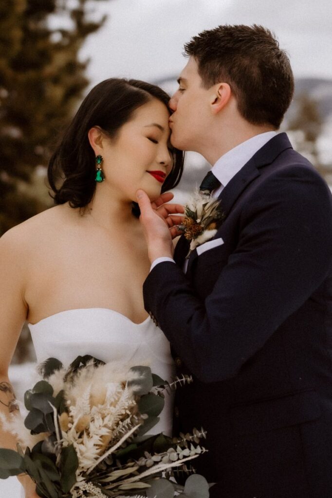 a man in a black suit holding the chin of his partner and kissing her forehead the woman is wearing a white wedding dress and holding flowers and smiling with her eyes closed 