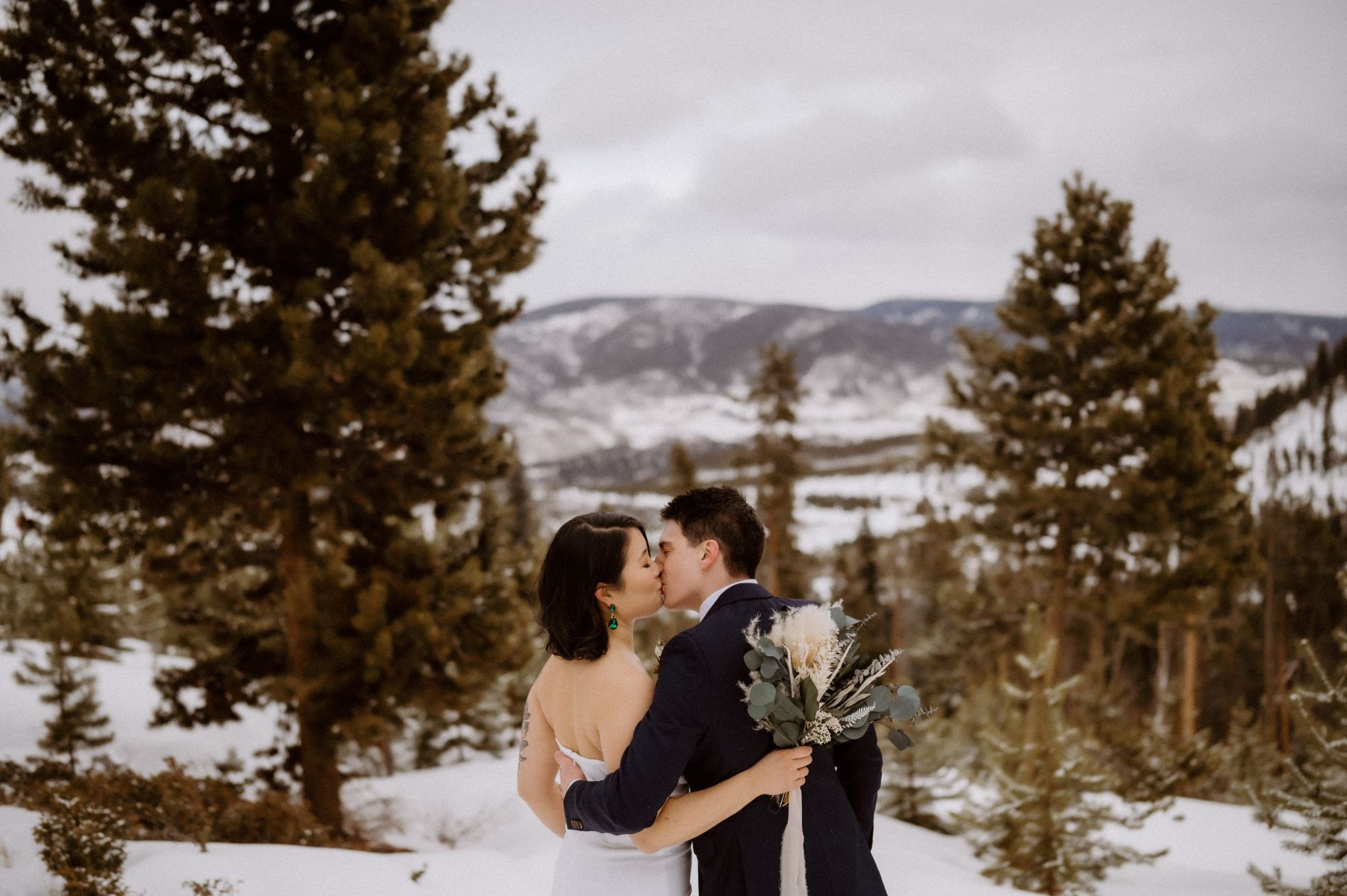 a couple kissing on top of an overlook at Sapphire Point in the winter in Colorado the woman is wearing a white wedding dress the man is wearing a black suit 