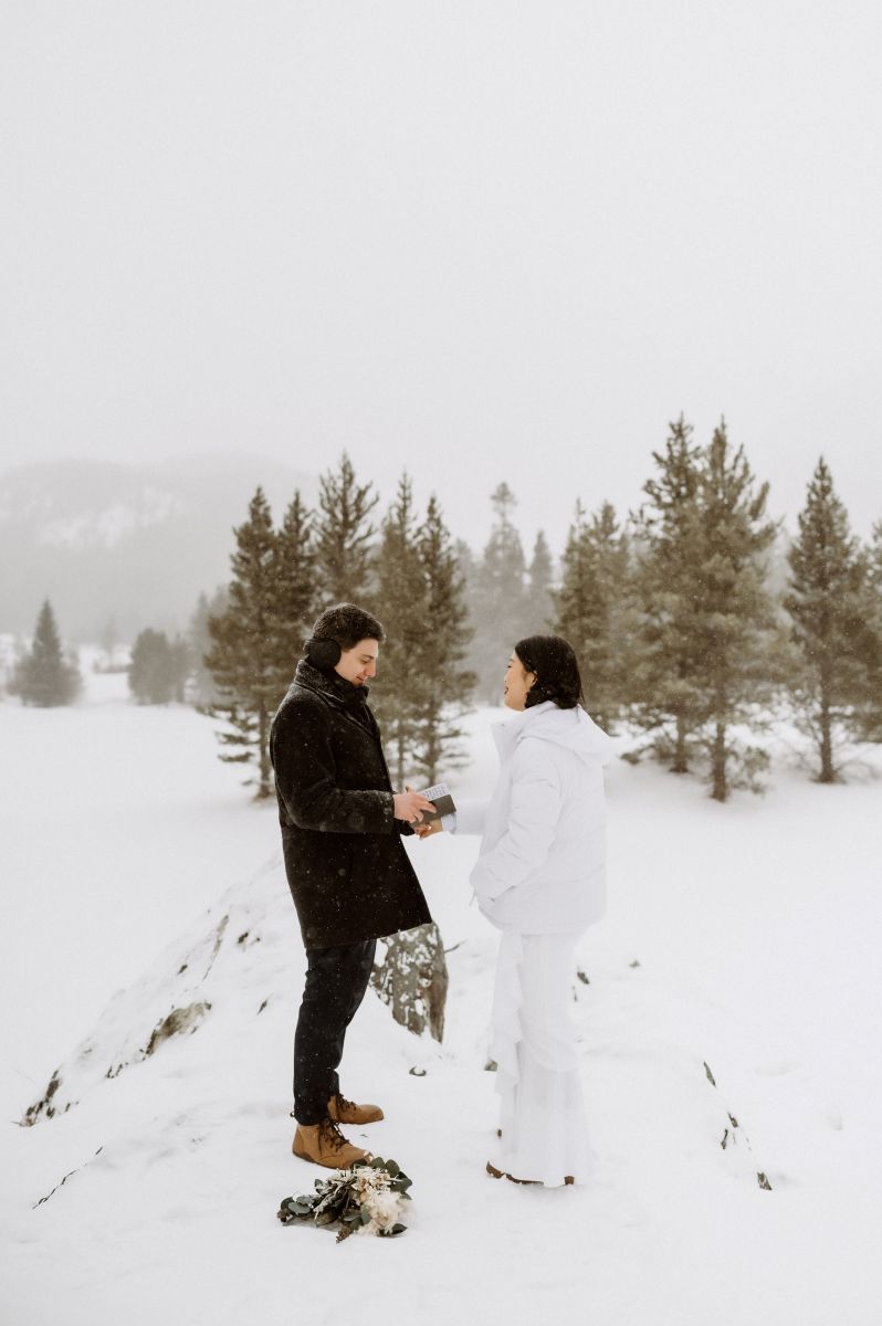 a couple exchanging vows in the snow the man is holding a vow book the woman is wearing a white jacket and wearing a white wedding dress the man is wearing a brown jacket and earmuffs 