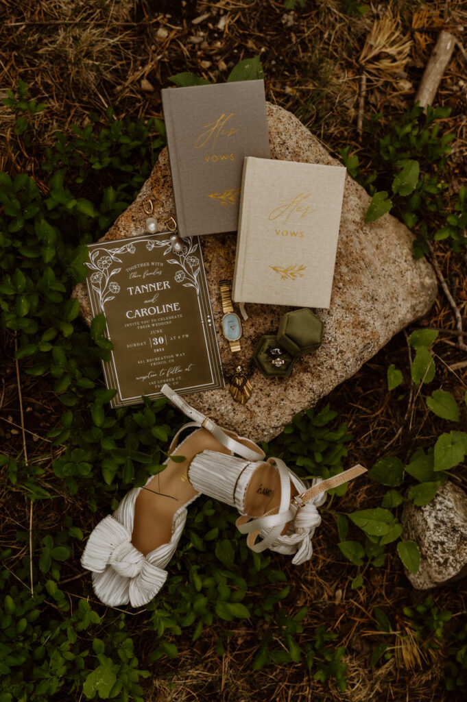 Wedding invitations, vow books, jewelry, and shoes laid on a rock in the woods 