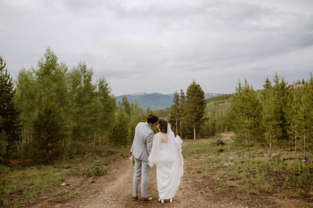 A newlywed couple holding hands and kissing while standing on a trail in the mountains 